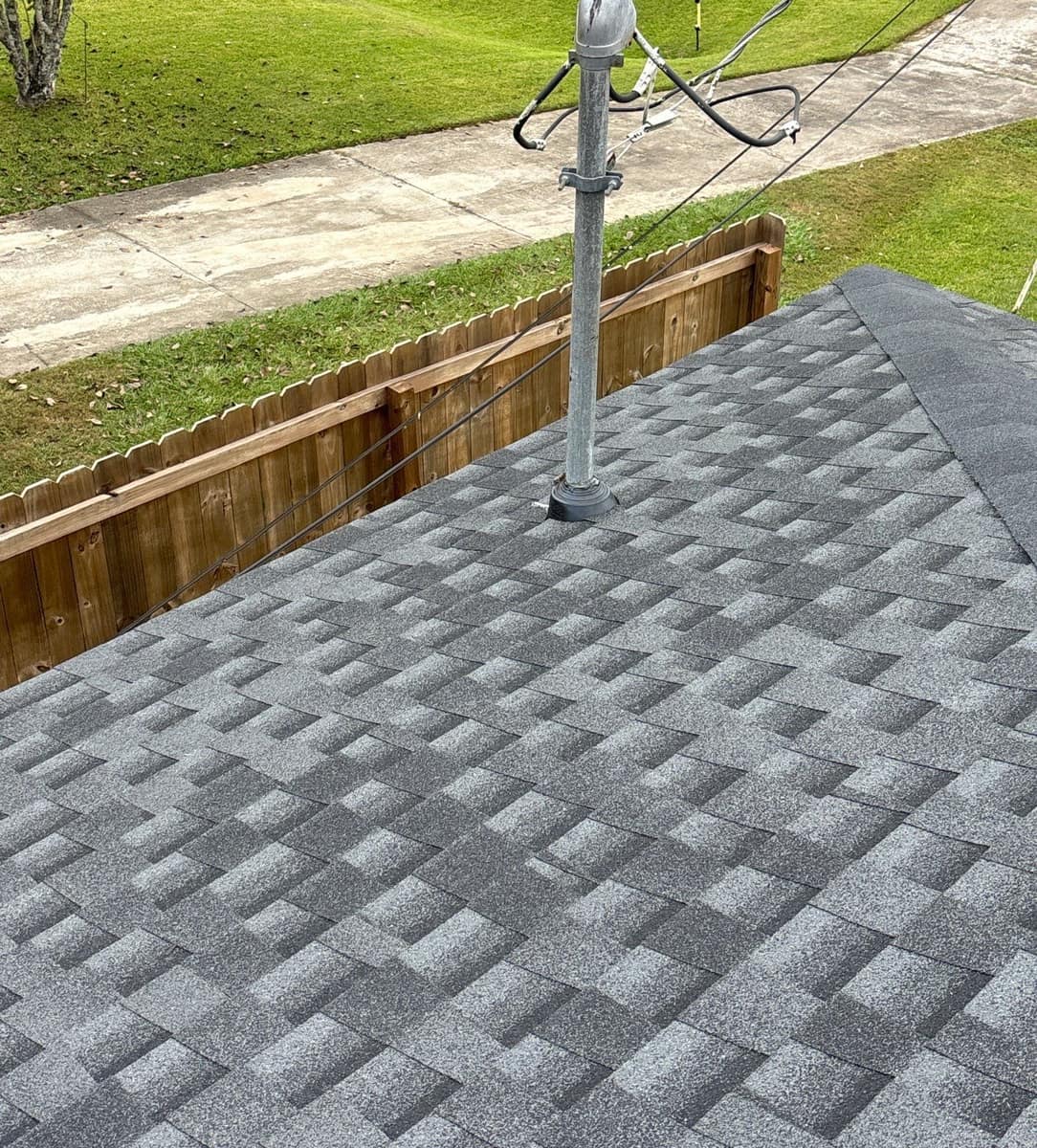 Electric service mast rising through the new shingles above a wood fence and lawn in Alachua