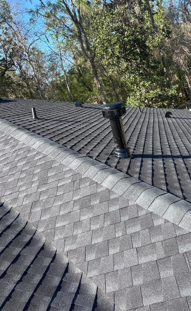 A tall black vent stack beside a capped hip, with woods beyond the Alachua roof