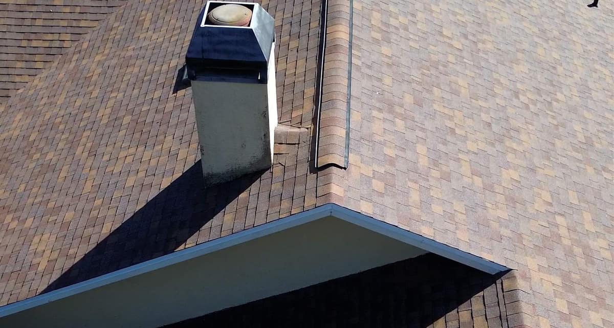 Stucco chimney with a dark metal cap rising through new shingles above a white gable eave in Alachua
