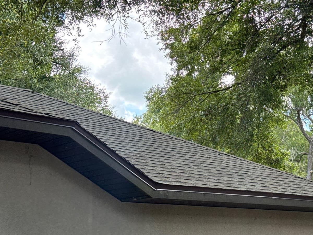 Looking up at a shingled hip corner and brown eave trim under oak branches in Gainesville