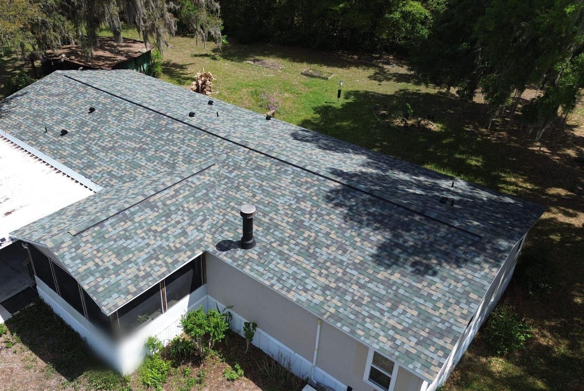 Drone view of a Reddick manufactured home with new multi-tone shingles and an older white metal carport roof
